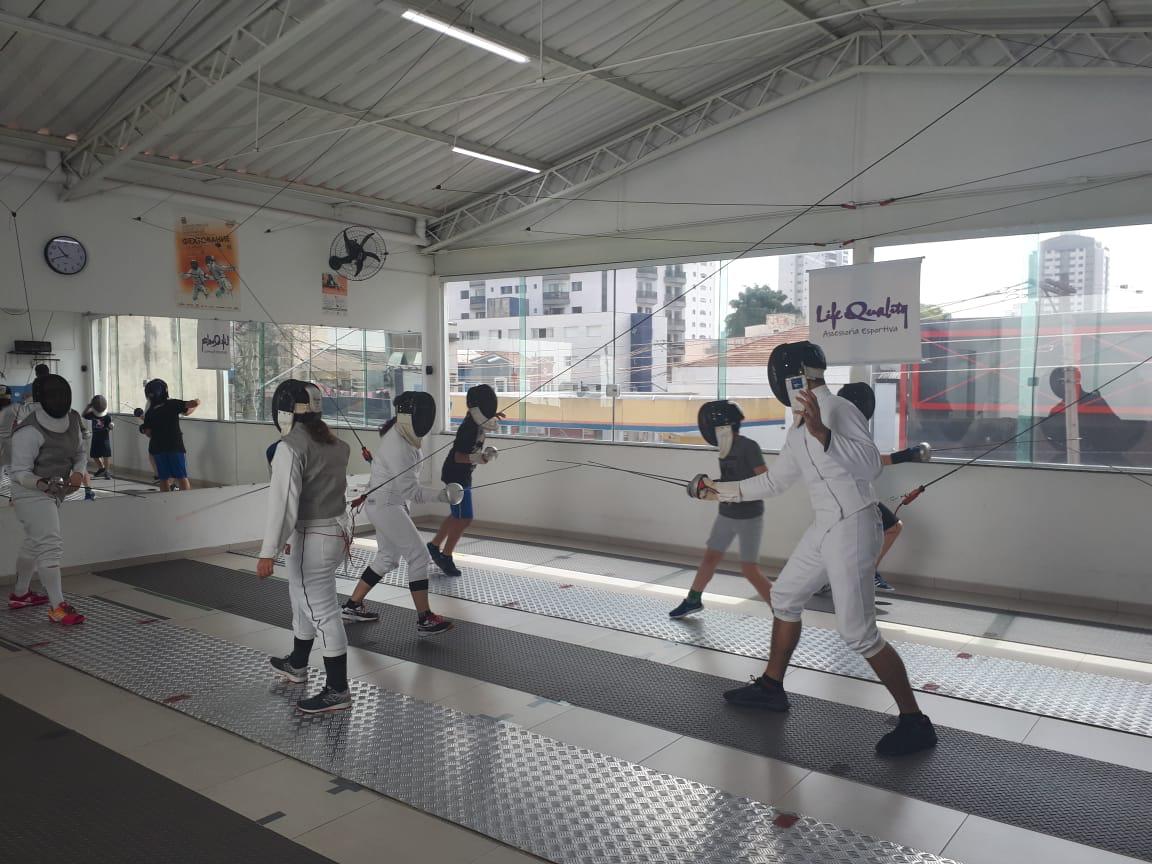 Alunos com vestimentas adequadas praticam esgrima em um ambiente interno, eles estão com espadas nas mãos, máscara próprias e uniformes brancos 